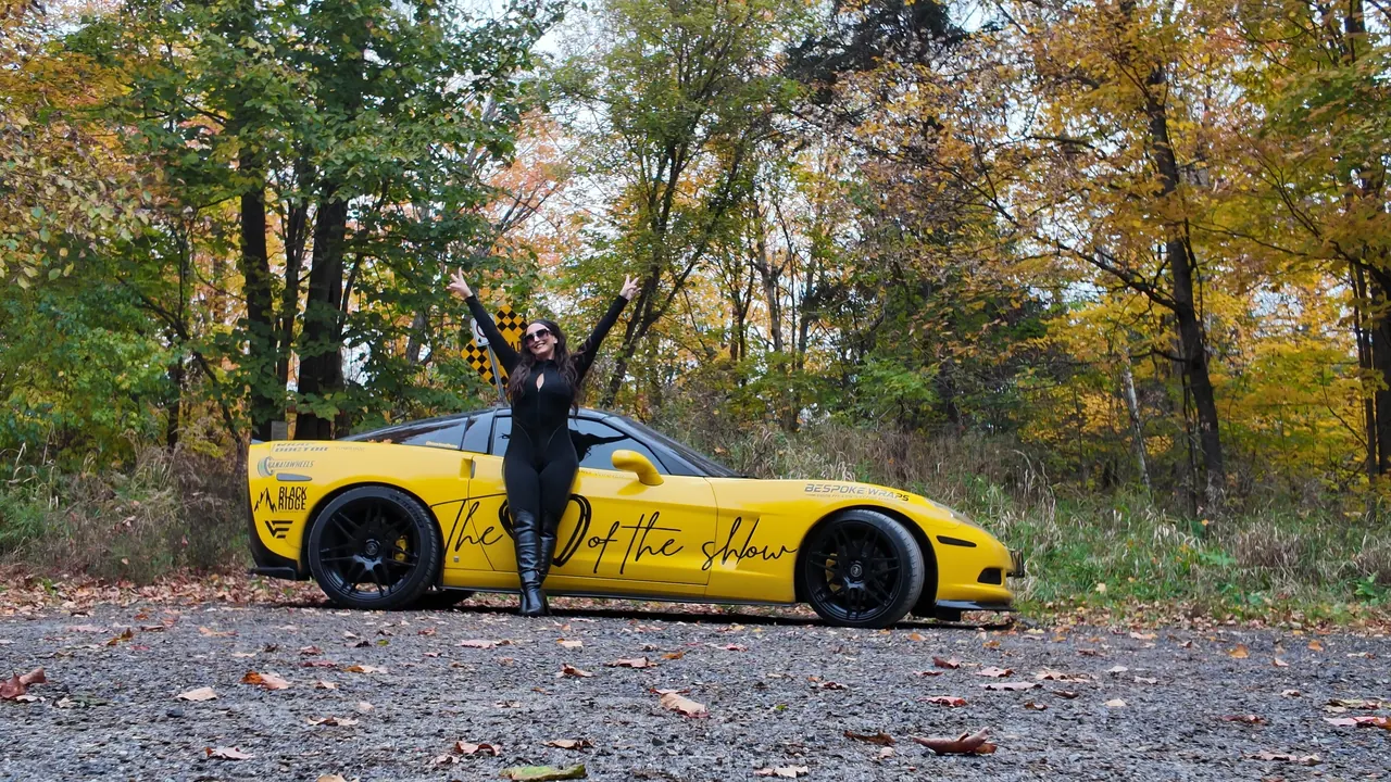 Chelsea with both arms raised in celebration in front of the Corvette.