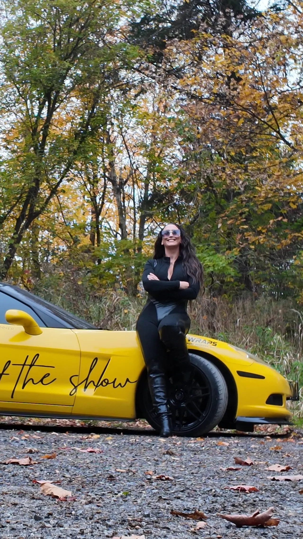 Chelsea Walsh leaning against her yellow Corvette, arms crossed, looking up toward the trees.