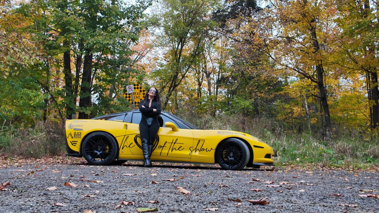 Chelsea standing with arms crossed beside the full length of her yellow Corvette.
