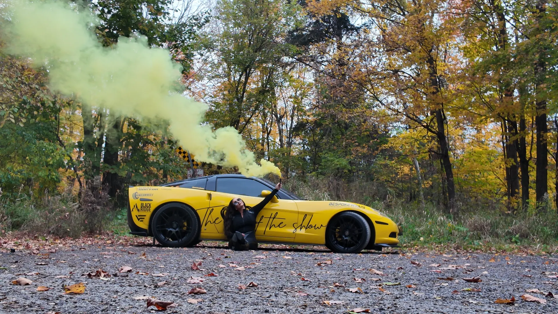 Chelsea Walsh crouched beside her yellow Corvette C6 as yellow smoke drifts into the autumn trees.