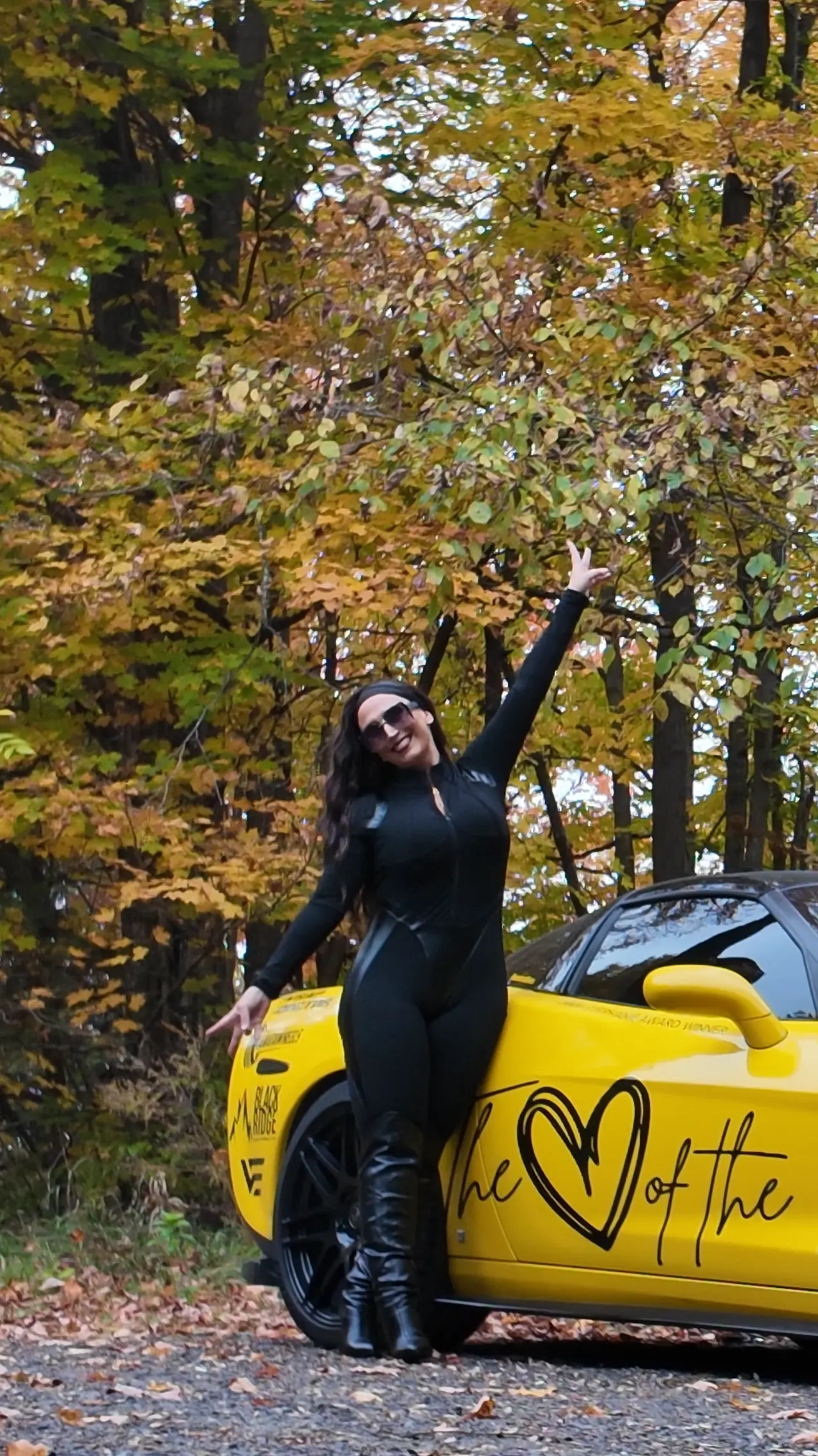 Chelsea waving with one arm raised beside the Corvette.